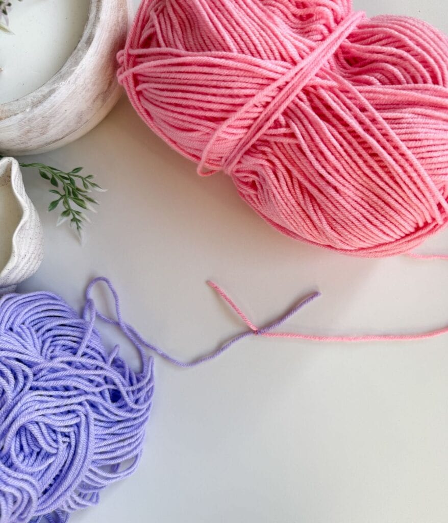 A ball of pink yarn and a loose pile of purple yarn rest on a white surface, ready to demonstrate how to join new yarn in crochet, with some green leaves and ceramic dishes partially visible in the corners.