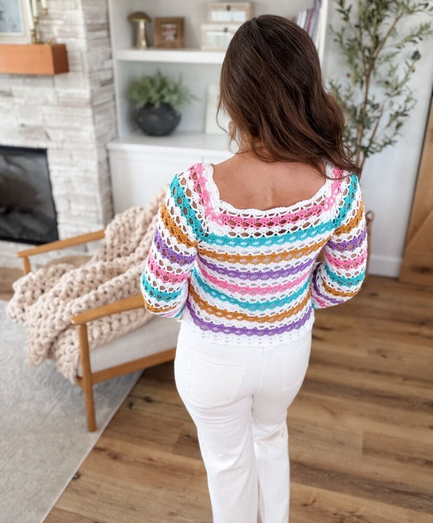 A person stands indoors facing away, wearing a colorful crocheted top featuring a delicate shell stitch and white pants, in a living room with wooden floors, a chair, and shelves with decor.