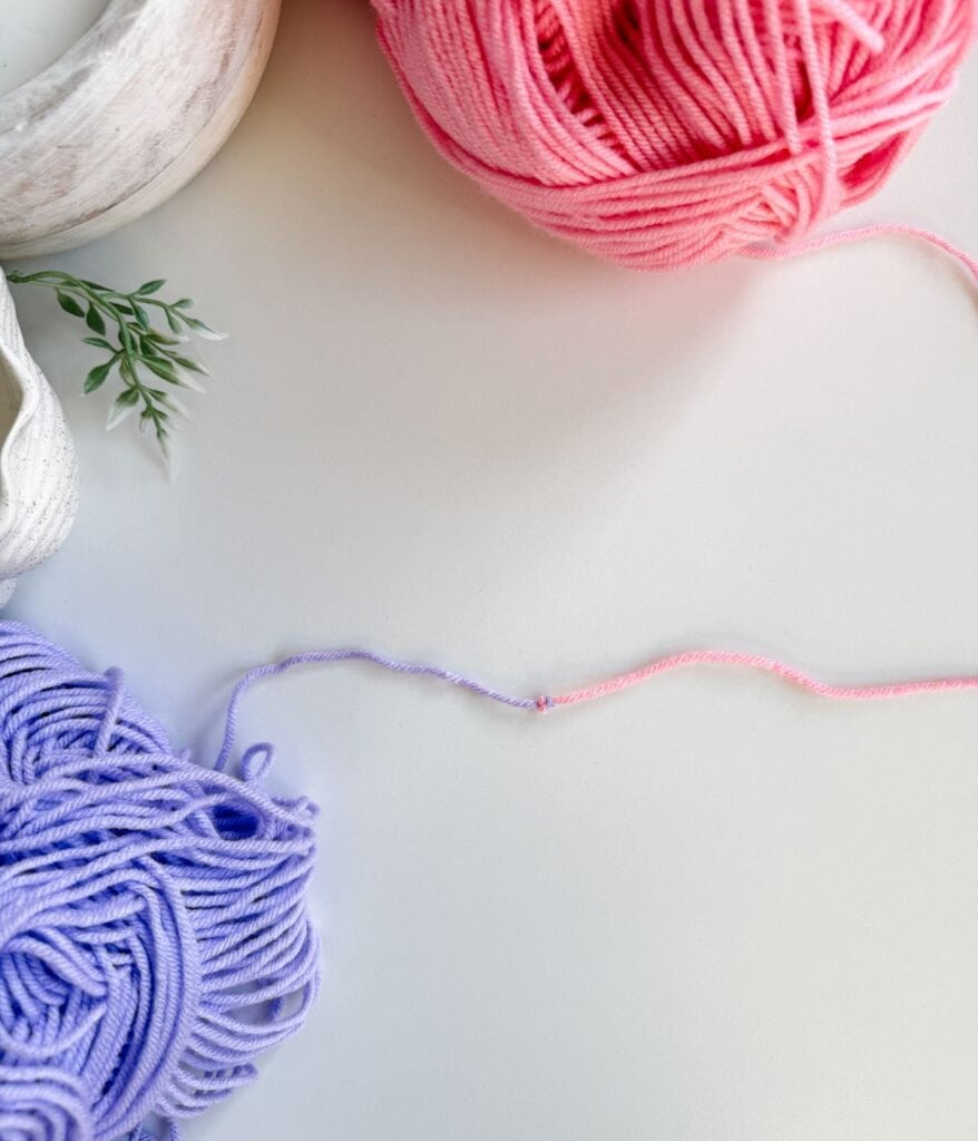 Pink and purple yarn balls sit on a white surface, with a loose strand connecting the two—a perfect illustration of how to join new yarn in crochet. A small plant and part of a white container are visible in the corner, completing this creative setup showcasing essential crochet techniques.