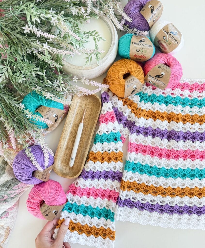 A multicolored crochet sweater is displayed with a crochet hook, wooden tray, yarn balls in various colors, and greenery above, showcasing a beautiful shell stitch design—perfect inspiration if you’re looking for a new crochet pattern or even searching for a free pattern to try.