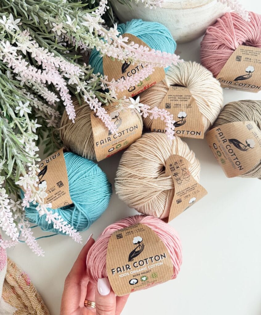 Six balls of Fair Cotton yarn in blue, cream, and pink are displayed on a white surface with lavender flowers above them. In the foreground, a hand holds one ball of pink yarnโperfect for your next crochet vest project.