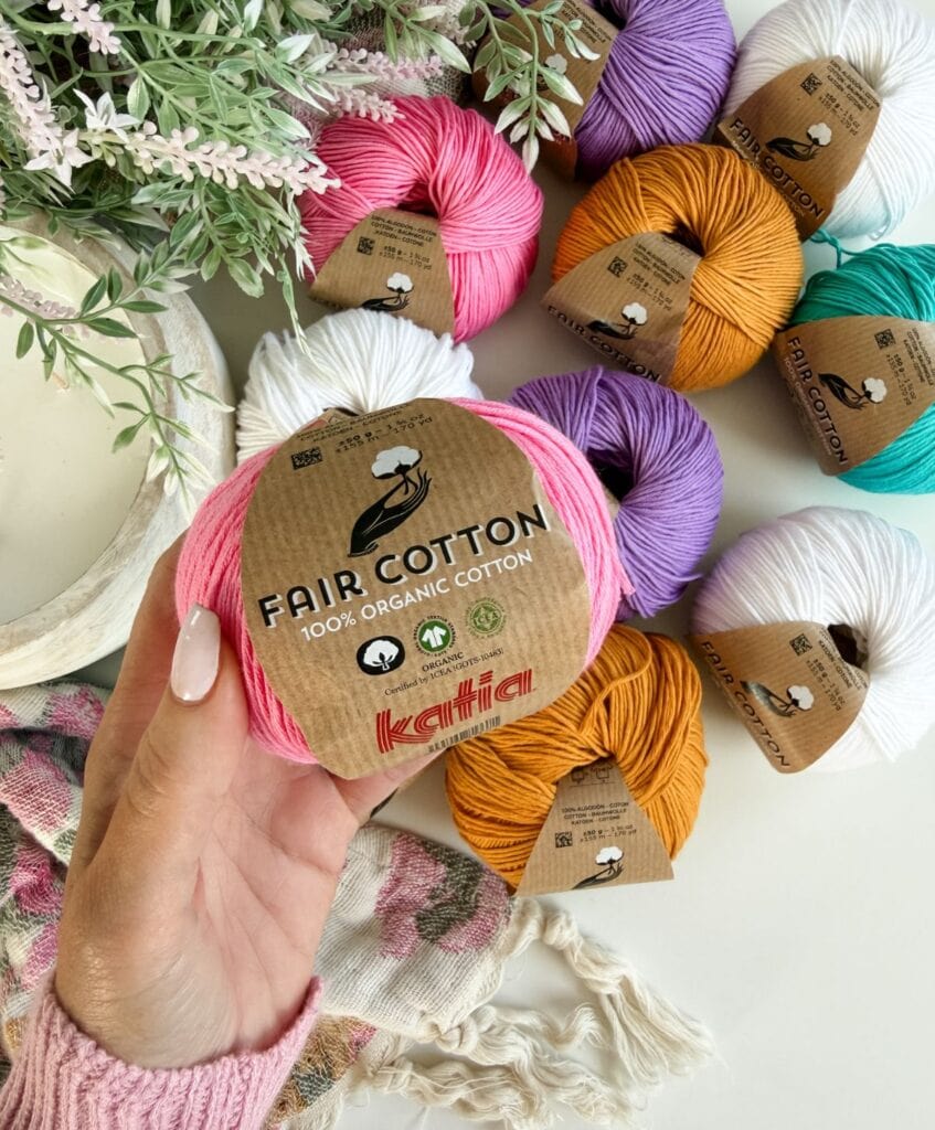 A hand holds a pink skein of Katia Fair Cotton yarn, perfect for your next Serena Shell Stitch project; various colored skeins and a plant are visible in the background, offering inspiration for your crochet pattern creations.