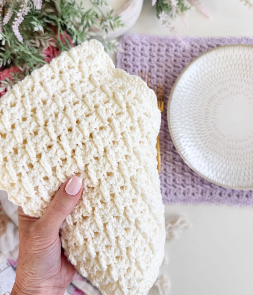 A hand holding a textured, cream-colored crocheted cloth next to a white plate on a light purple crochet placemat pattern, with greenery in the background.