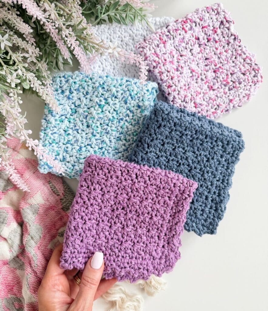Five textured crocheted squares in various colors, including purple, blue, gray, and multicolored, are displayed on a white surface with decorative foliage in the background. Each piece beautifully showcases an easy crochet dishcloth pattern perfect for adding both style and functionality to your handmade collection.
