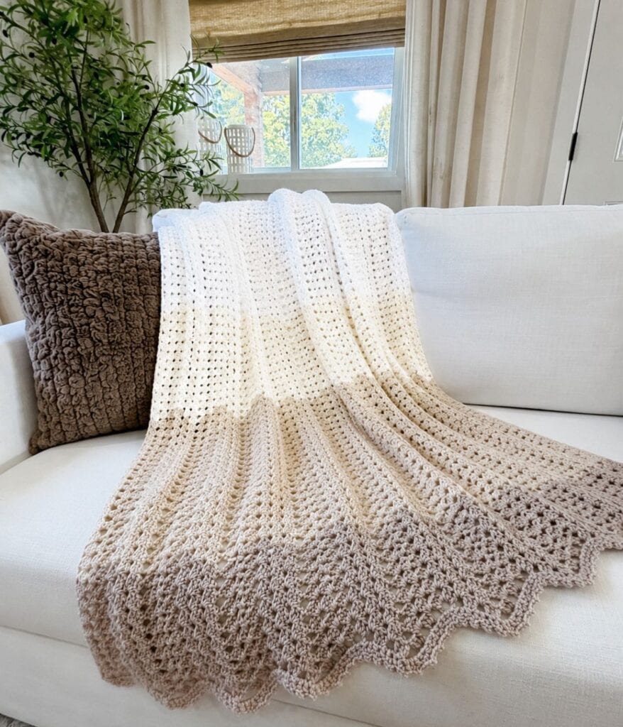 A beige and cream crochet blanket, crafted using a classic granny ripple blanket crochet pattern, is draped over a white sofa with a brown textured pillow, near a window with greenery outside.