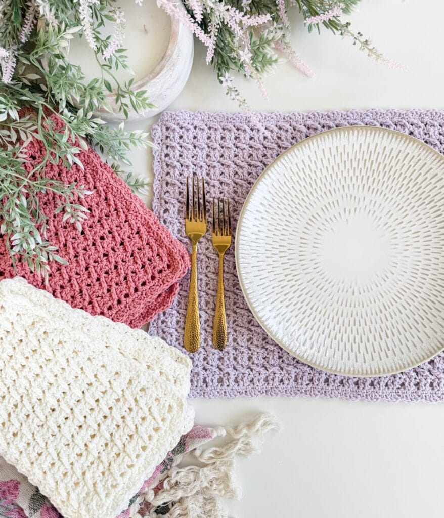 A white plate and two gold forks are placed on a lavender crocheted placemat, made using a delicate crochet placemat pattern, with folded pink, cream, and lavender crochet pieces nearby and leafy plants above.