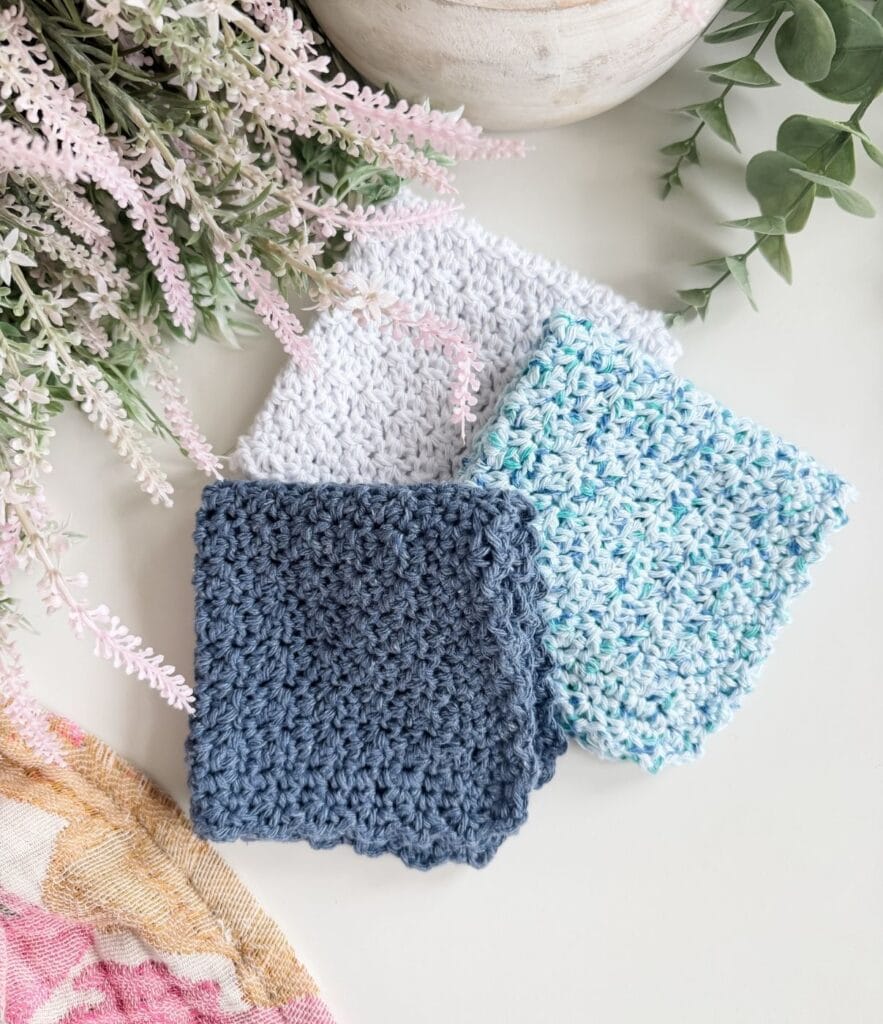 Three crocheted square washcloths in blue, white, and variegated blue yarn—made using an easy crochet dishcloth pattern—are arranged on a white surface near green and pink leafy plants.