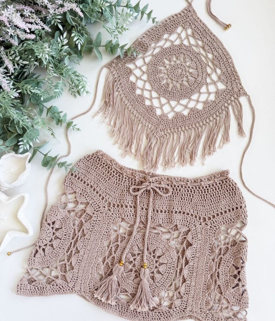 Two beige crochet garments lay flat: a fringed halter top with a square mandala design, and a matching skirt with tassels and intricate patterns. Perfect as a boho resort set, these pieces feature an eye-catching crochet pattern. Greenery and seashell decor are nearby.
