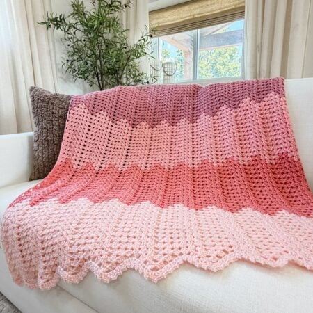 A pink ombre granny ripple blanket crochet pattern drapes elegantly over a white couch, complemented by pillows and a potted plant in the background.