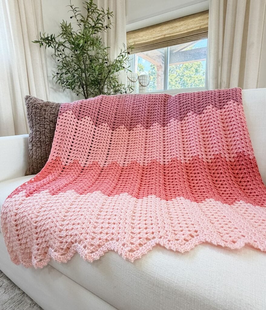 A pink ombre granny ripple blanket crochet pattern drapes elegantly over a white couch, complemented by pillows and a potted plant in the background.