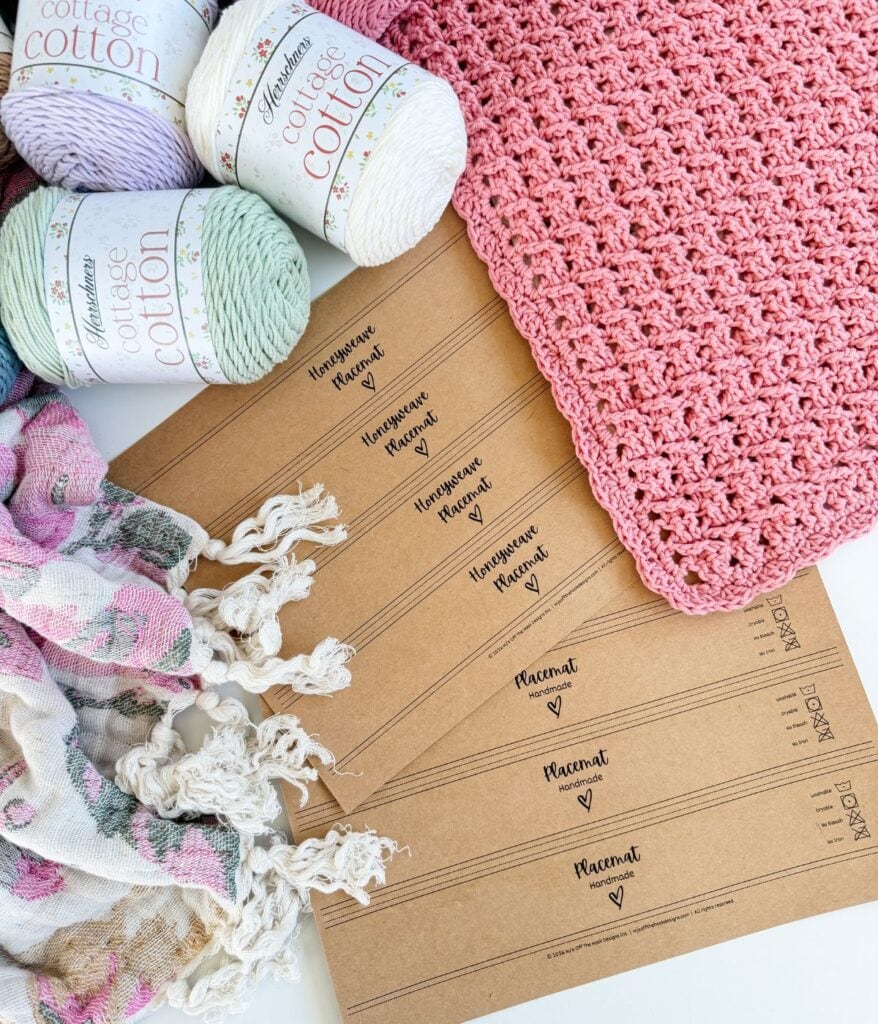Pink crocheted fabric, yarn skeins, a multicolored scarf with tassels, and five brown labels reading “Handmade with love, Placental” are arranged on a white surface, offering inspiration for your next crochet placemat pattern.