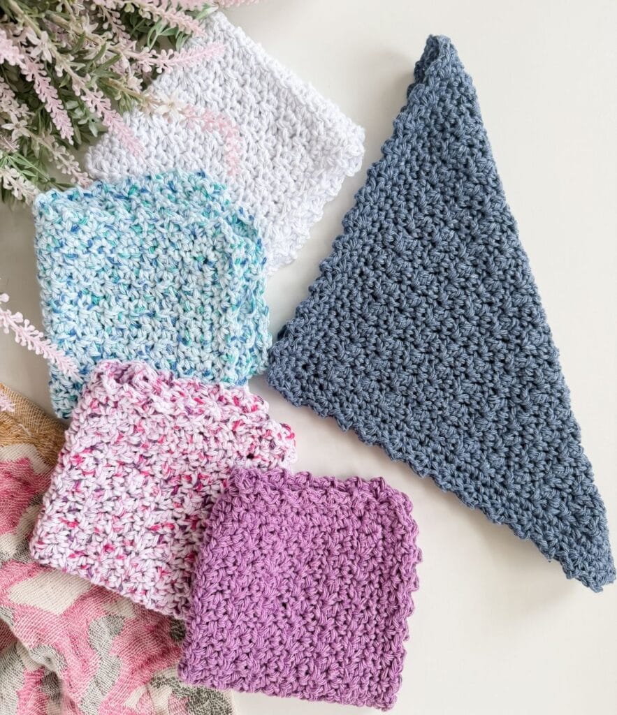 Five crocheted swatches in various colors and shapes are arranged on a white surface, showcasing an easy crochet dishcloth pattern, with lavender flowers partially visible in the background.