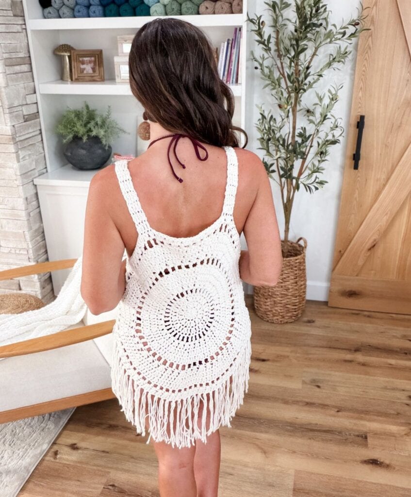 A woman with long brown hair wearing a white crocheted dress with fringe stands indoors, facing away from the camera. The intricate crochet pattern of her dress highlights the delicate craftsmanship, reminiscent of the timeless elegance found in Solana Circle designs.