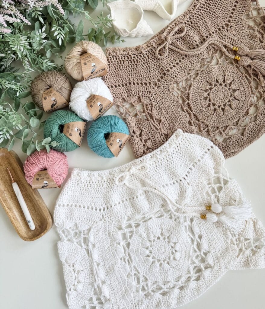 Two crocheted tops, one beige and one white, from the Zahara Boho Resort Set, are displayed with balls of yarn, a crochet hook, and green leaves on a white surface—a perfect showcase of this unique crochet pattern.