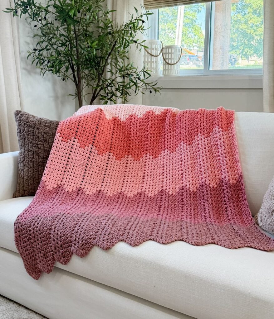 A pink ombre granny ripple blanket crochet pattern, featuring a zigzag design, is draped over a white couch next to brown pillows in a bright living room.