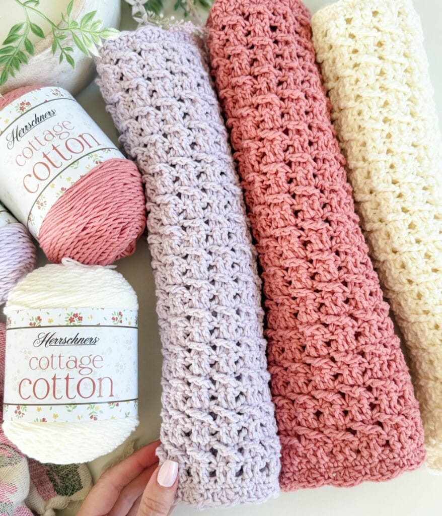 Three rolled crochet blankets in lavender, pink, and cream colors rest beside skeins of Herrschners Cottage Cotton yarn on a light surface—perfect inspiration for your next crochet placemat pattern.