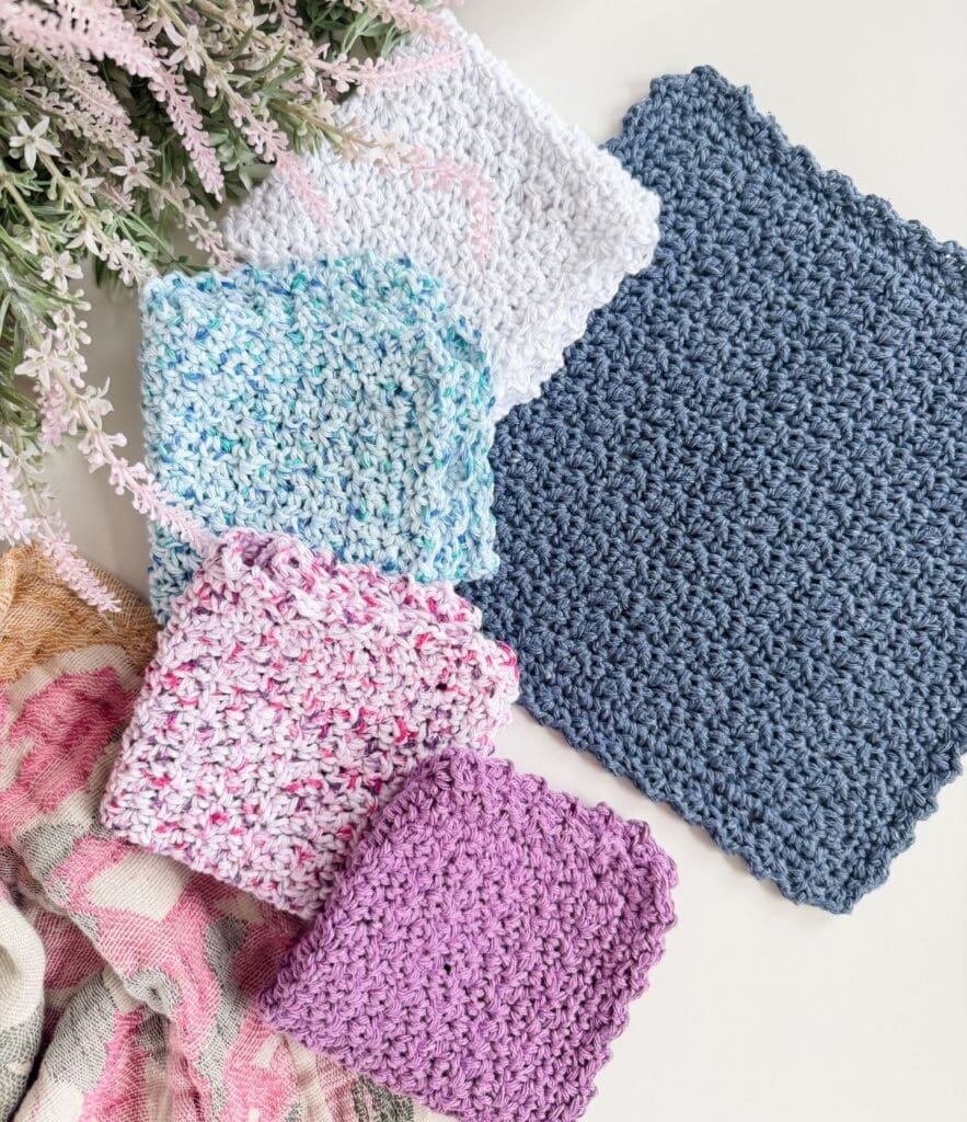 Five crocheted square cloths in various colors are arranged on a white surface next to pink and green artificial flowers and a patterned fabric, showcasing an easy crochet dishcloth pattern perfect for beginners or adding a handmade touch to your kitchen.