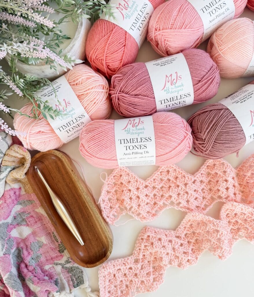 Six skeins of pink yarn, a wooden yarn bowl, a partially crocheted pink zigzag piece reminiscent of a granny ripple blanket crochet pattern, and a floral fabric arranged on a white surface with lavender flowers.