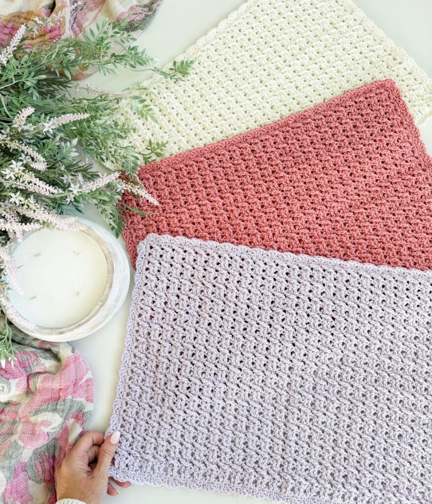 Three textured, crocheted placemats in cream, pink, and lavender are laid out on a table next to a candle, greenery, and a floral cloth. A hand holds the lavender placemat’s corner, showcasing the intricate crochet placemat pattern that adds charm to any table setting.