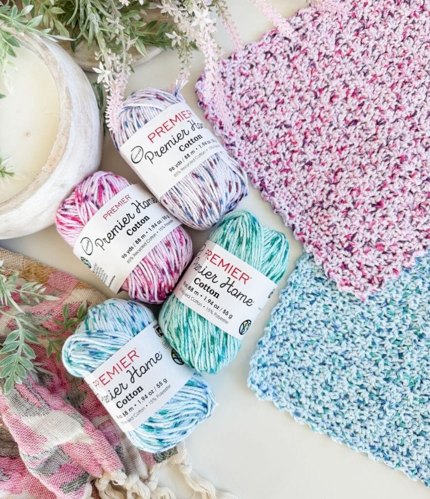 Four skeins of Premier Home Cotton yarn are arranged next to two knitted swatches—perfect for sampling an easy crochet dishcloth pattern—alongside a candle, greenery, and a pink plaid cloth on a white surface.