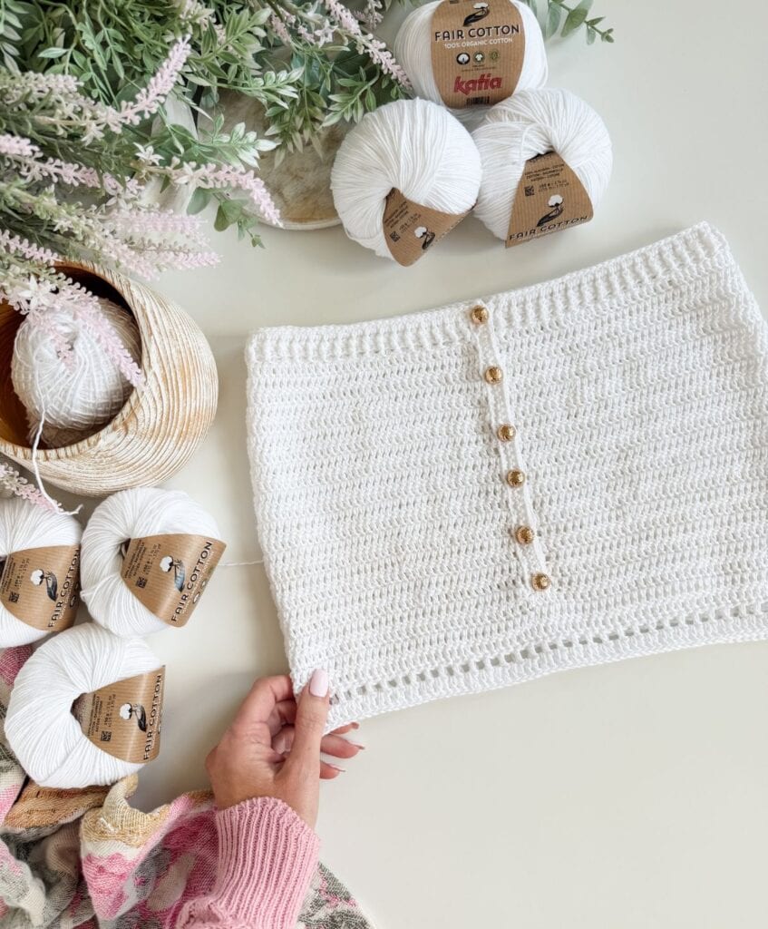 A hand holds a white crocheted skirt with buttons, surrounded by white yarn balls, a pink sweater sleeve, and green foliage on a white surface—perfect inspiration for your next Crochet Beach Dress or to try out a Free Crochet Beach Dress Pattern.