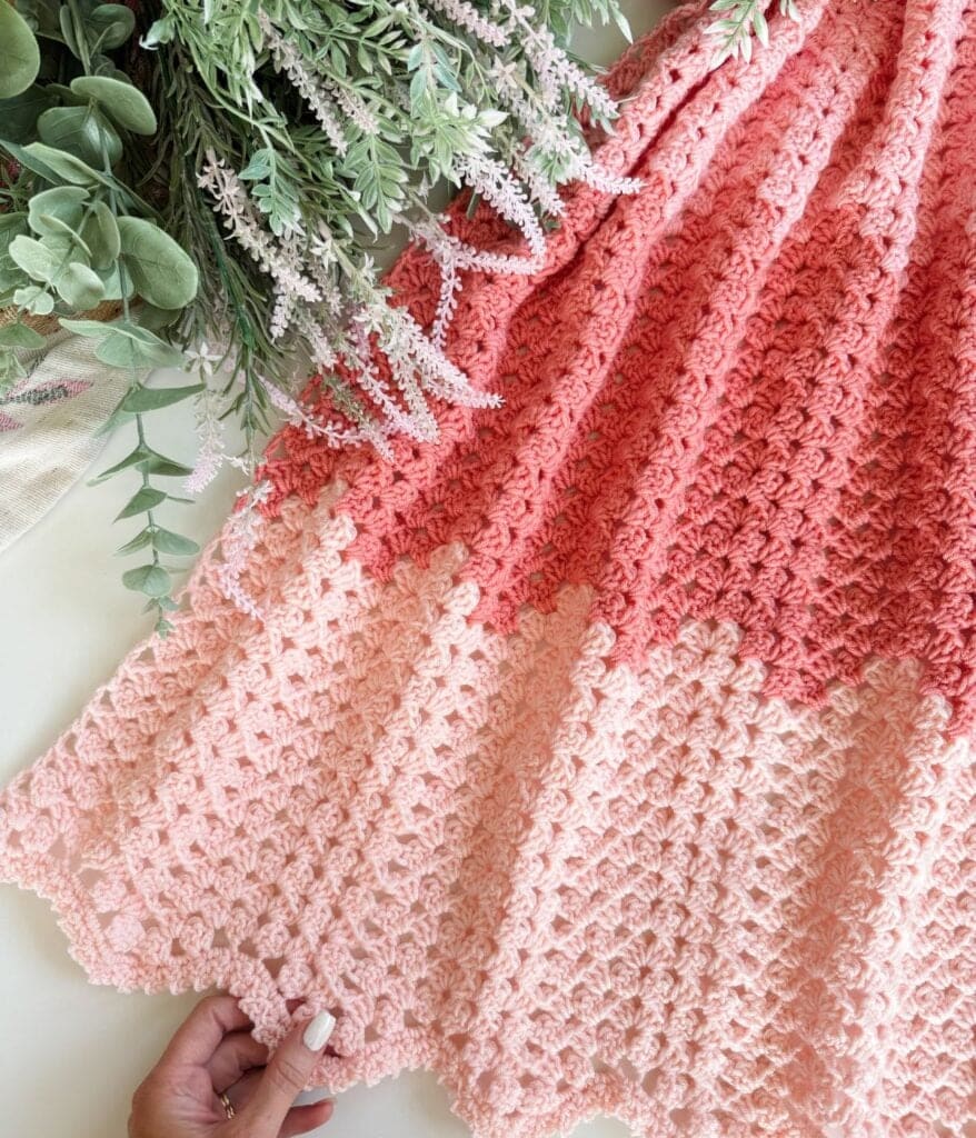 A hand holds a pink and peach granny ripple blanket crochet pattern with a textured design, next to a bunch of green and purple artificial foliage.