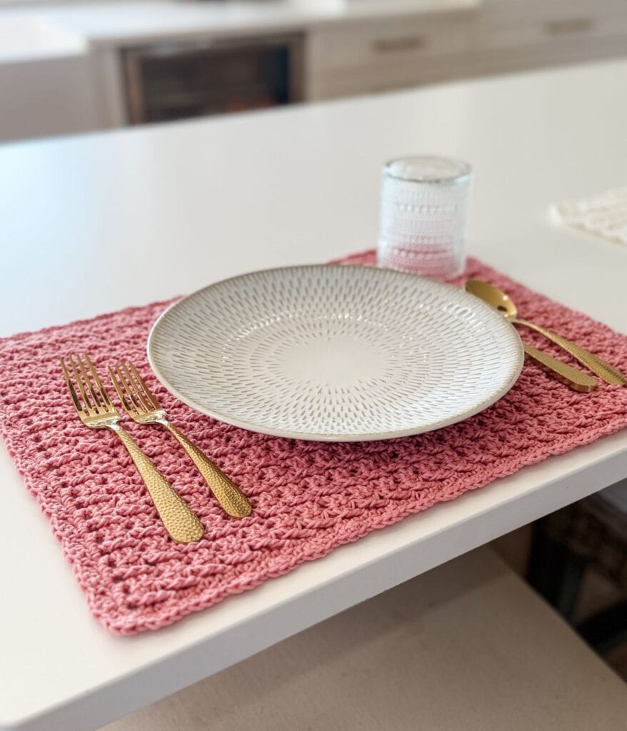 A white patterned plate, gold cutlery, and a clear glass are arranged on a pink crocheted placemat—crafted with an intricate crochet placemat pattern—set atop a white table.
