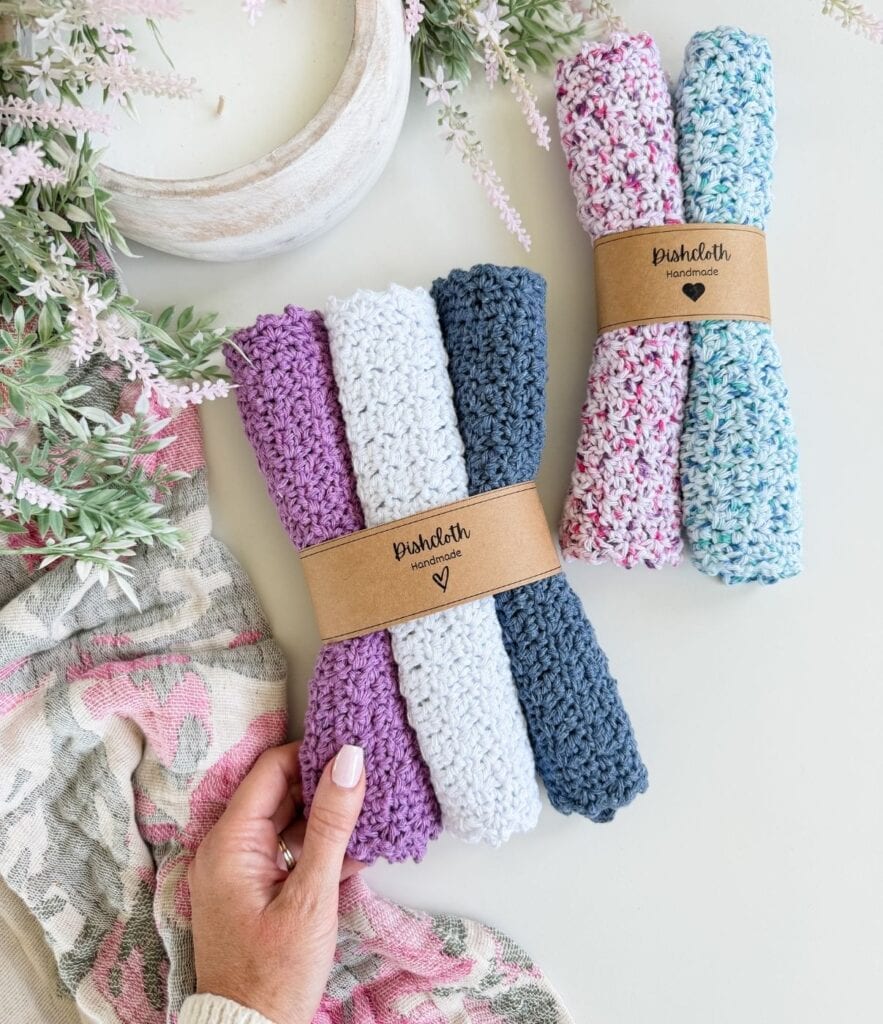 Hand holding a set of three knitted dishcloths in purple, white, and blue, wrapped with a brown label; another set in pink, white, and blue—crafted with an easy crochet dishcloth pattern—is displayed on a white surface with greenery nearby.