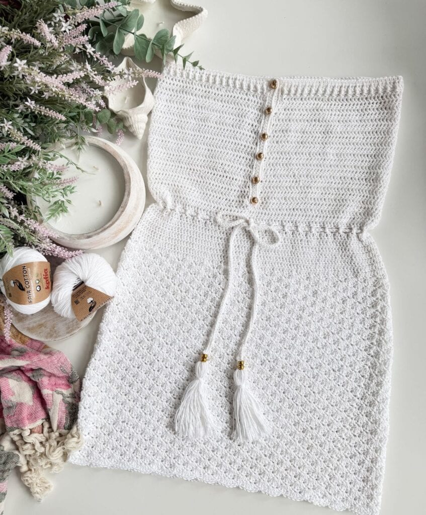 A handmade white crocheted dress, inspired by the Saltwater Shell Beach Dress, with front buttons and tassel drawstrings is displayed alongside yarn, crochet hooks, and floral decorations on a white surface.