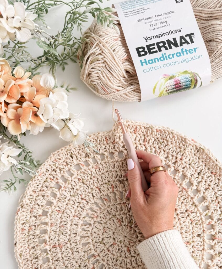 A hand crochets a round beige piece using the Solana Circle Crochet Pattern next to a skein of Bernat Handicrafter cotton yarn and a bunch of pale artificial flowers on a white surface.