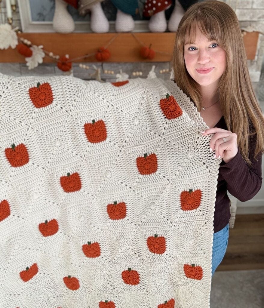 A woman stands indoors holding a cream-colored crocheted blanket decorated with orange pumpkin squares, showcasing a charming crochet pumpkin blanket pattern.