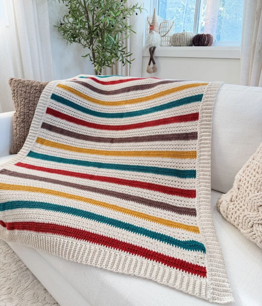 A crochet blanket pattern featuring red, yellow, teal, and brown stripes is draped over a white sofa in a bright, cozy living room.