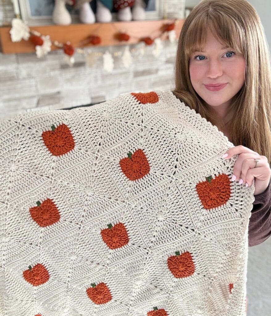 A woman holds a handmade crochet pumpkin blanket, featuring a cream background and orange pumpkin patterns, standing indoors near a decorated fireplace mantel. This cozy scene beautifully showcases the crochet pumpkin blanket pattern in an inviting autumn setting.