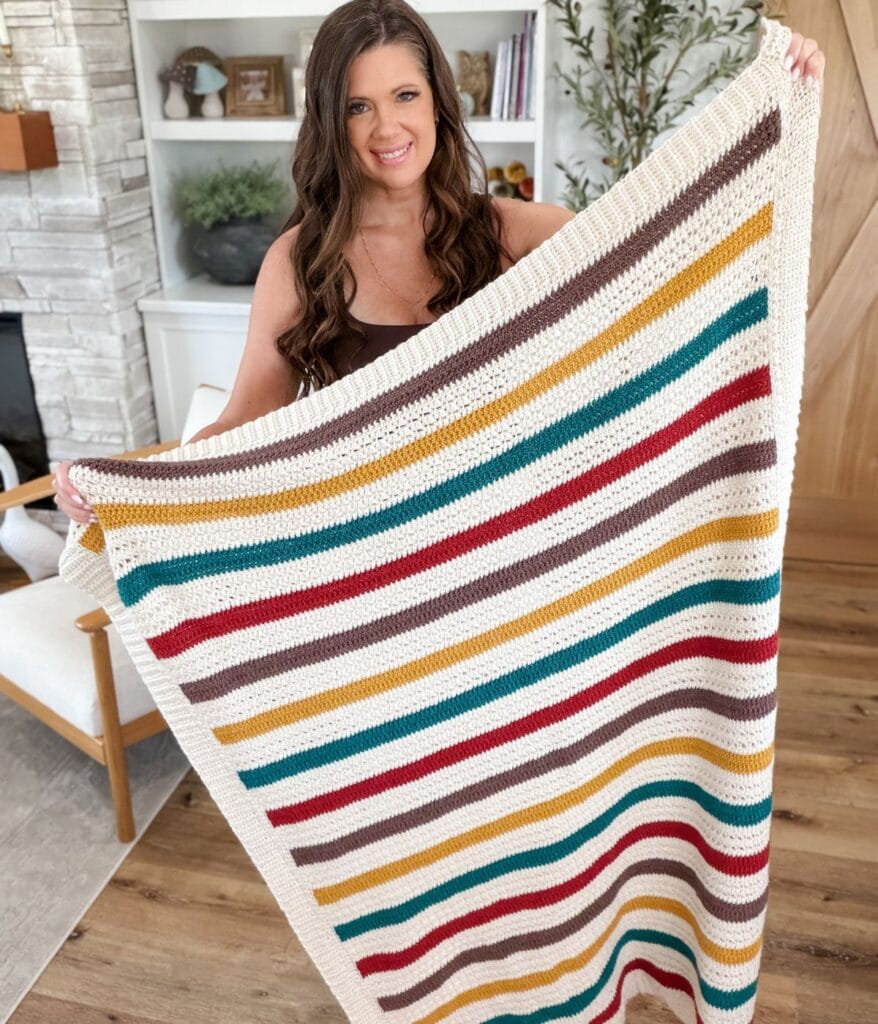 Woman holding a crocheted blanket with horizontal stripes in teal, mustard, red, and brown—crafted from a beautiful crochet blanket pattern—standing in a living room with wood floors and neutral decor.