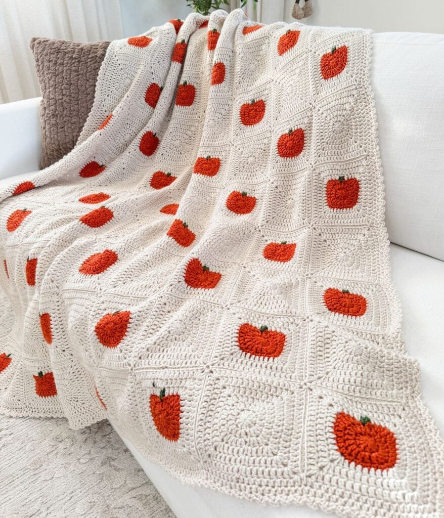 Beige crocheted blanket with red and green apple motifs, inspired by a crochet pumpkin blanket pattern, draped over a white couch and taupe textured pillow in a bright room.