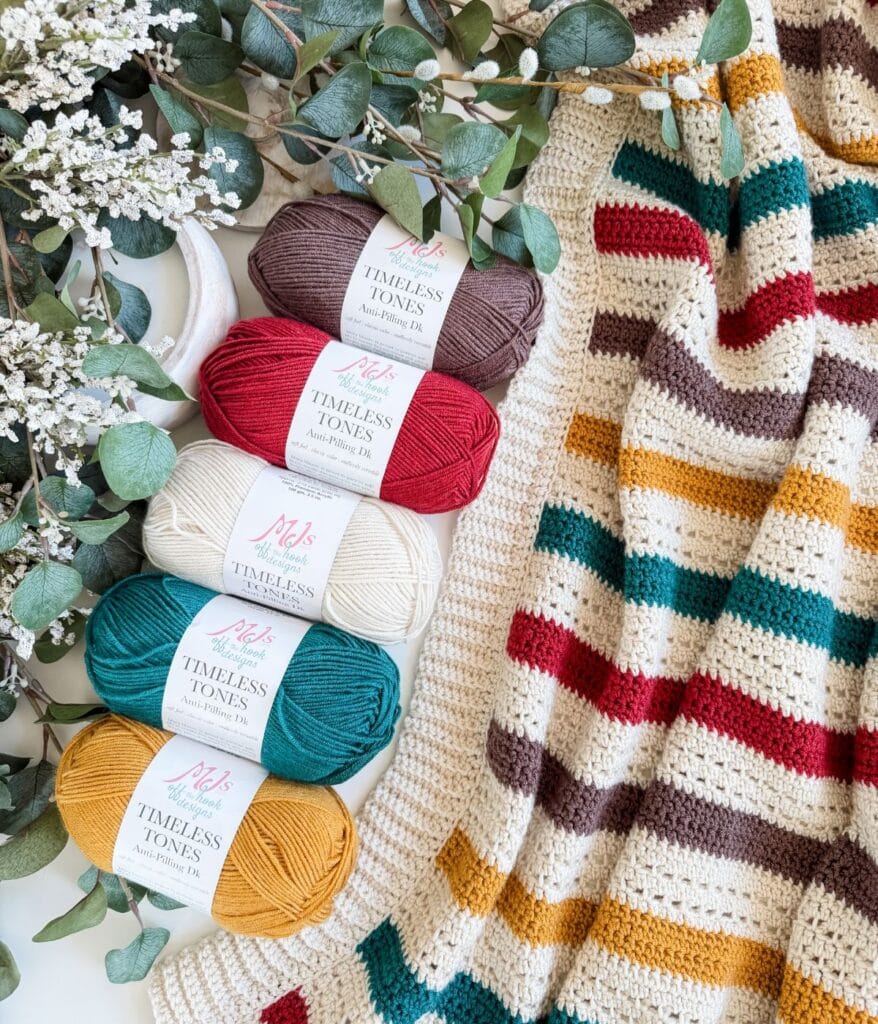 Four skeins of yarn in gold, teal, cream, and burgundy sit beside a striped crochet blanket and green eucalyptus branches with white flowers, inviting you to try a new crochet blanket pattern.