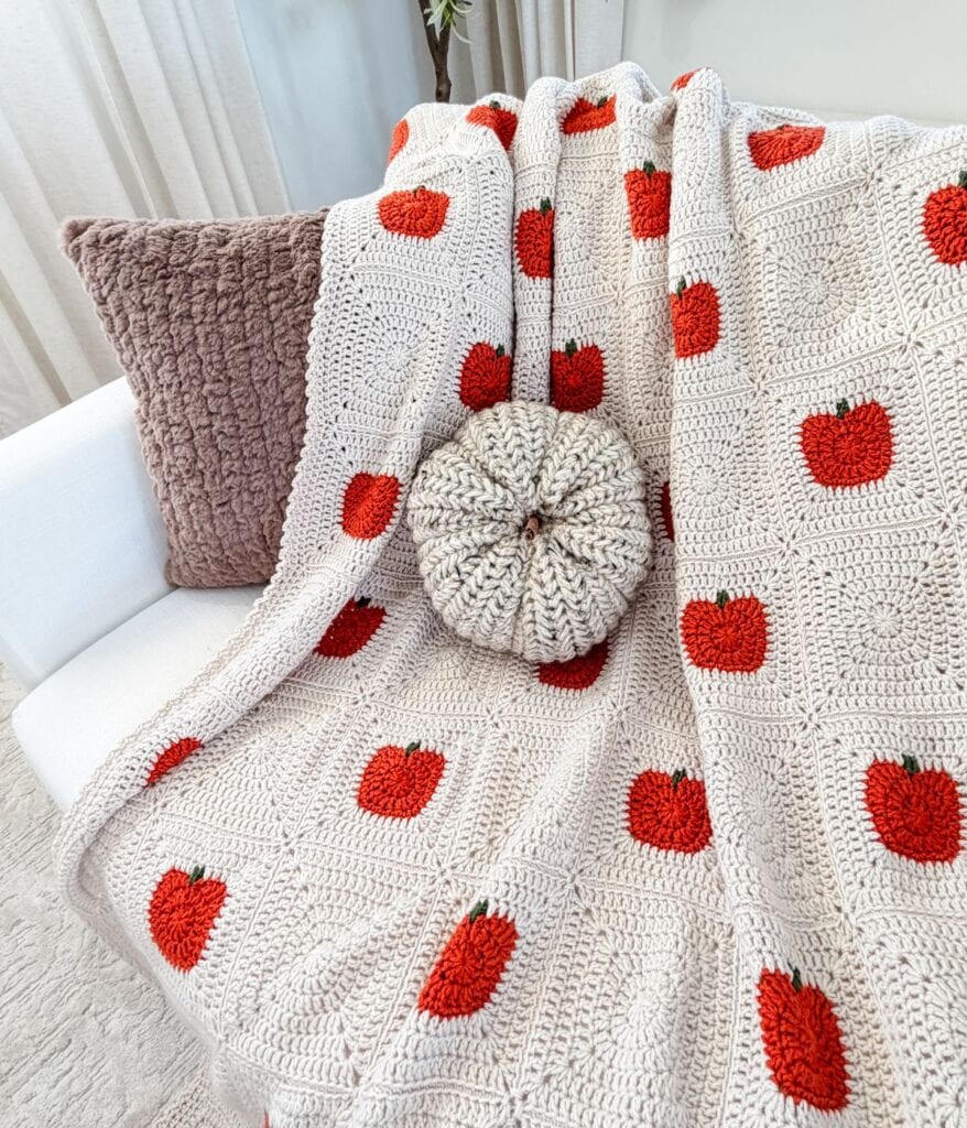 A crocheted blanket with red apple patterns, inspired by a crochet pumpkin blanket pattern, is draped over a white chair, next to a textured brown pillow and a round, knitted cushion.