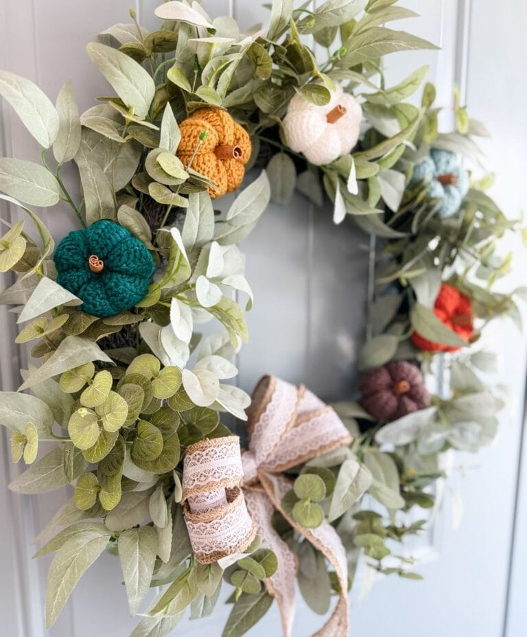 A wreath of green leaves decorated with small pumpkins made using the easiest pumpkin crochet pattern in various colors and a lace ribbon bow, hanging on a door.