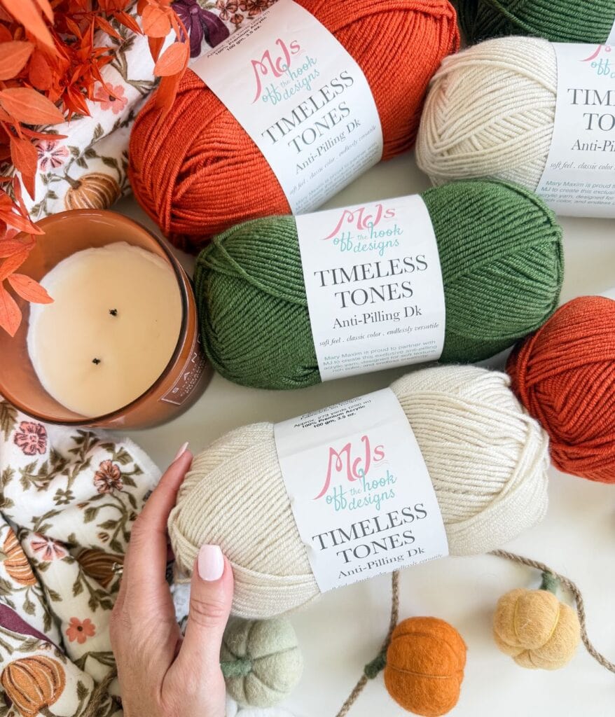 Three skeins of yarn in orange, green, and cream labeled “Timeless Tones” are arranged with a candle, felted balls, and autumn decor on a floral fabric—perfect for your next crochet pumpkin blanket pattern. A hand touches the cream yarn.