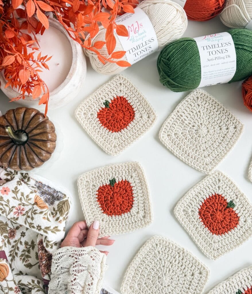 Five crocheted squares with orange pumpkin designs are laid out on a surface surrounded by yarn, a faux pumpkin, orange foliage, and a person’s hand holding one square—perfect inspiration for anyone looking to try a new crochet pumpkin blanket pattern.