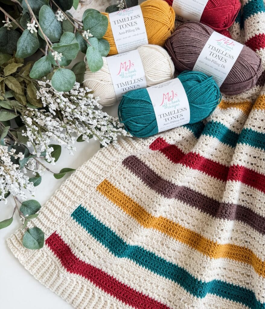 A striped crocheted blanket, made using a charming crochet blanket pattern, lies next to sprigs of eucalyptus and balls of colorful yarn labeled “Timeless Tones.”.