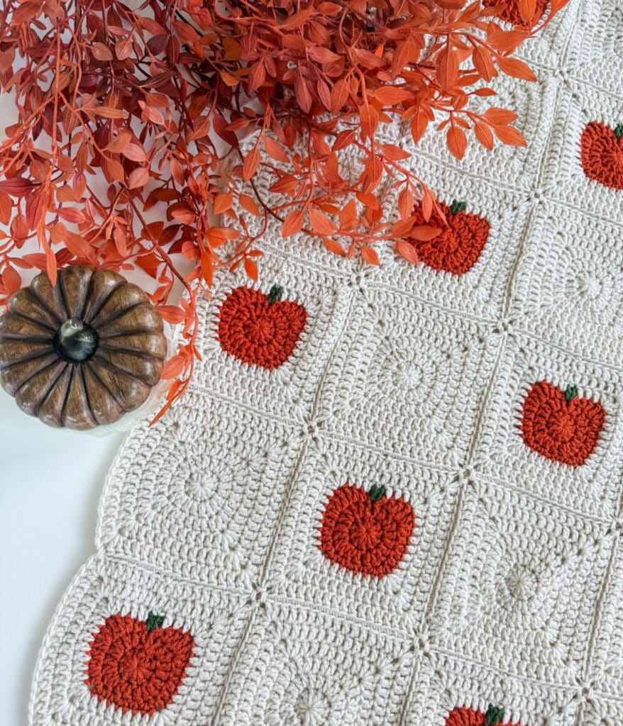 Crocheted white blanket with red apple patterns, next to an artificial brown pumpkin and orange leaves on a white surface—perfect for pairing with your favorite crochet pumpkin blanket pattern for a cozy, autumn-inspired touch.