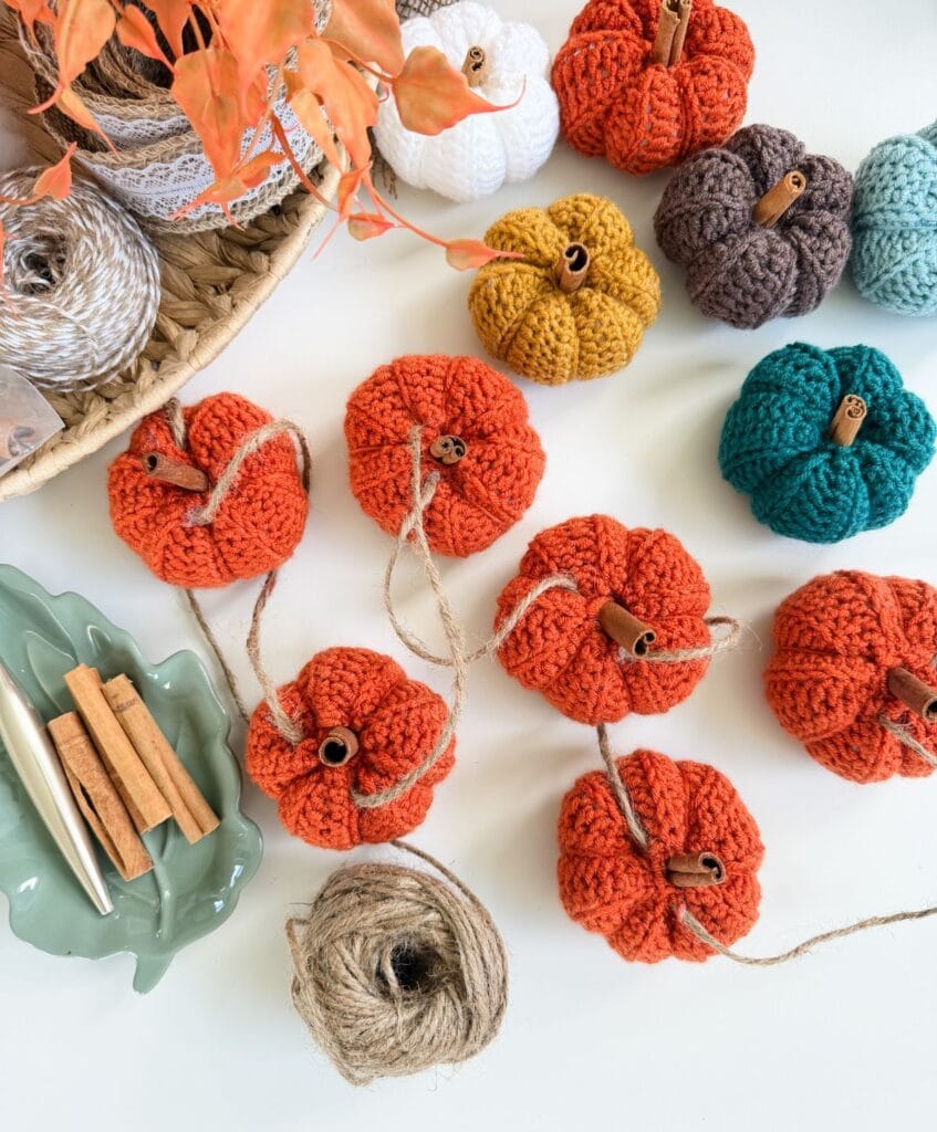 Crocheted pumpkins in various colors are arranged on a table with twine, cinnamon sticks, and crochet tools nearby, crafted using the easiest pumpkin crochet pattern for charming autumn decor.