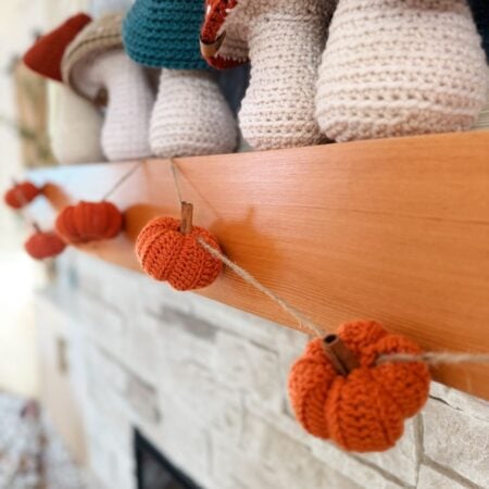 Crocheted orange pumpkins, made using the easiest pumpkin crochet pattern, hang from a twine garland draped across a wooden mantel, with knitted decor items displayed above on the shelf.