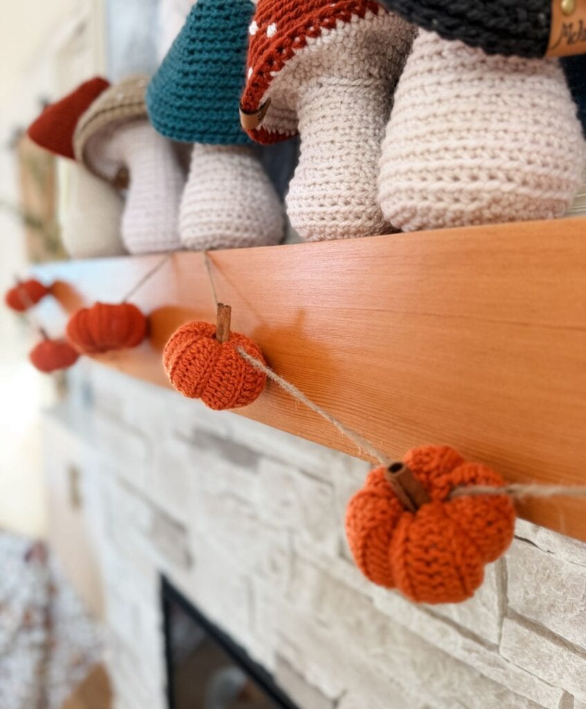 Crocheted orange pumpkins, made using the easiest pumpkin crochet pattern, hang from a twine garland draped across a wooden mantel, with knitted decor items displayed above on the shelf.