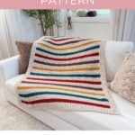 A striped crochet throw blanket with autumn colors, crafted from a beautiful crochet blanket pattern, is draped over a white sofa, with a textured beige pillow and a brown blanket beside it.