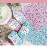 Three colorful crocheted dishcloths and four skeins of Premier yarn are displayed on a white surface with greenery on the side, promoting an easy crochet dishcloth pattern perfect for beginners.