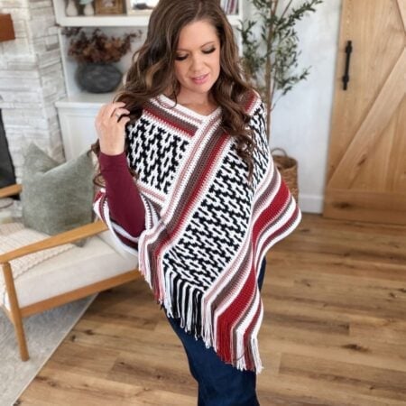 A person wears a black, white, and maroon mosaic crochet poncho with fringe trim over a maroon long-sleeved shirt and blue jeans.