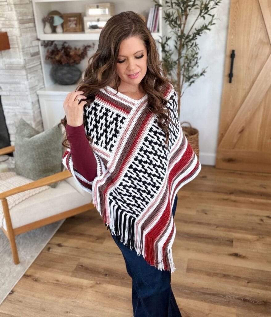 A person wears a black, white, and maroon mosaic crochet poncho with fringe trim over a maroon long-sleeved shirt and blue jeans.