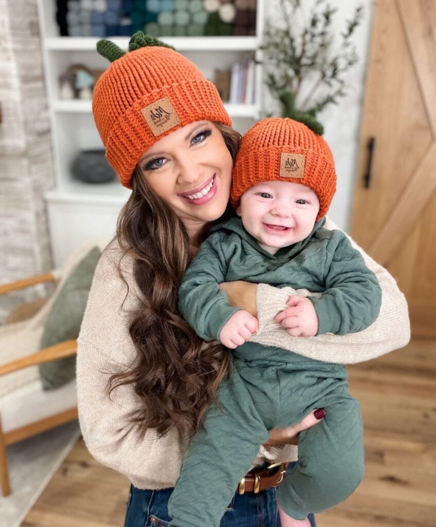 A woman and a baby wearing matching crochet pumpkin hats, with the baby smiling.